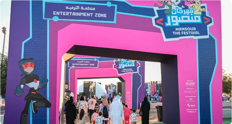 Daytime entrance gate with Mansour Festival branding and families entering the venue.